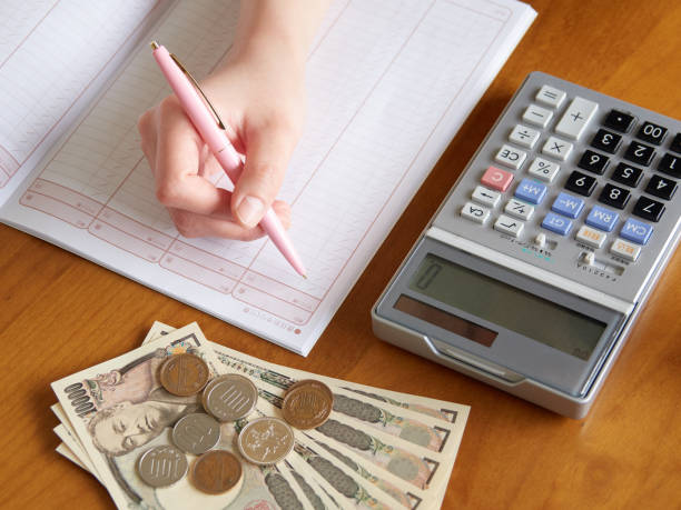 Person recording payroll figures with Japanese yen, coins, and a calculator on a desk
